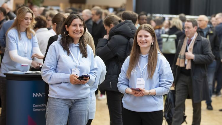 Two student assistants wearing light blue hoodies from the Northwest Alliance are standing in a large room full of people. 