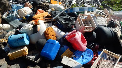 A pile of larger plastic pieces (broken beverage crates, a bobby car, lids from a trash can, baskets, canisters, pipes...) lies on dark volcanic stones.