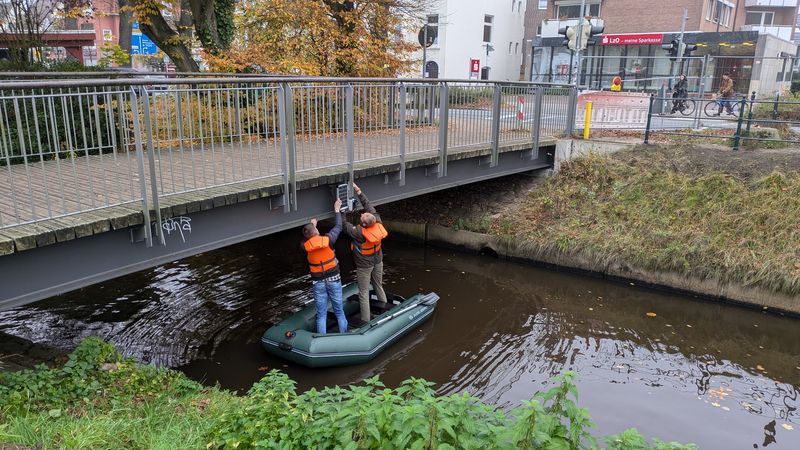 Zwei Masterstudierende bringen eine Sensorikbox zur Messung des Wasserspegels an einer Brücke an. Sie stehen dabei im Schlauchboot auf dem Fluss Haaren. Sie tragen eine orangene Rettungsweste.