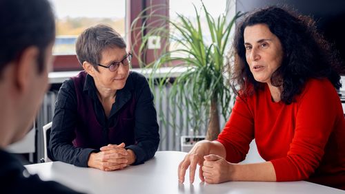 The interviewees sit opposite each other. Prof. Polat explains something to the interviewer while Iris Gereke looks at her from the side. The interview takes place in an office; a window and an indoor plant are situated in the background.