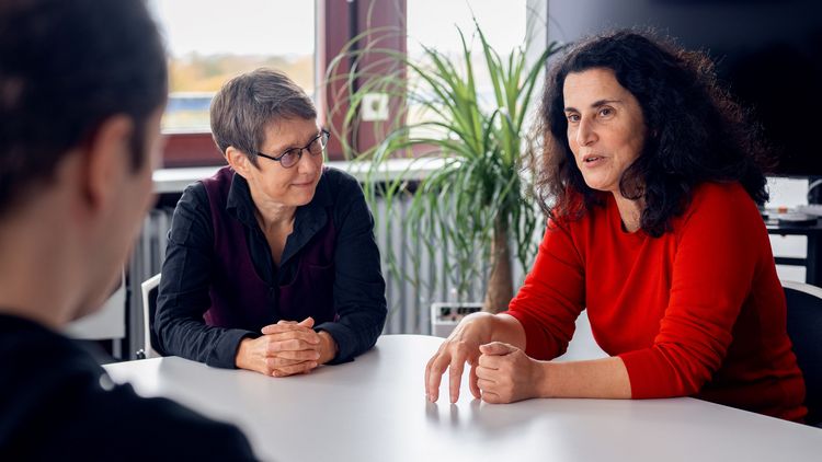 Die Gesprächspartner sitzen sich gegenüber. Frau Polat erklärt dem Interviewer einen Sachverhalt, während Frau Gereke sie von der Seite anschaut. Das Interview findet in einem Büro statt, im Hintergrund sind ein Fenster und eine Zimmerpflanze zu erkennen.