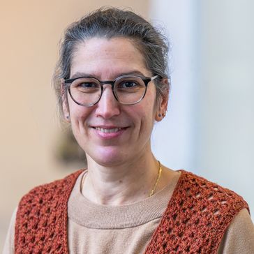 Frau mit beigem Pullover, dunklen Haaren, Brille und rot-gestricktem Pullunder lächelt in die Kamera. 