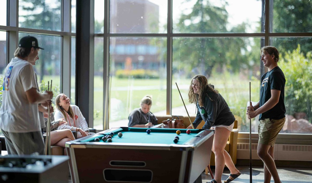 Studierende spielen Billard in einem hellen Aufenthaltsraum mit großen Fenstern.