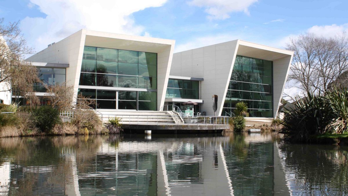 Ein moderner Gebäudekomplex der University of Waikato mit Glasfronten, der sich in einem See spiegelt.