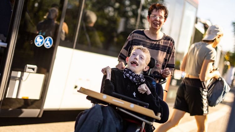 Das Bild zeigt einen jungen Mann, der unter einer schweren Beeinträchtigung leidet. Er sitzt in einem Rollstuhl. Er lacht sehr herzlich in die Kamera. Hinter ihm steht eine Frau und schiebt den Rollstuhl. Im Hintergrund sind ein Bus und eine Haltestelle sowie weitere Passagiere zu erkennen. Der junge Mann und die Frau sind gerade aus dem Bus ausgestiegen.