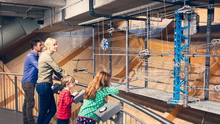 Eine vierköpfoge Familie steht an einem Gitter und probiert eine riesige Kugelbahn aus, die sich dahinter befindet.