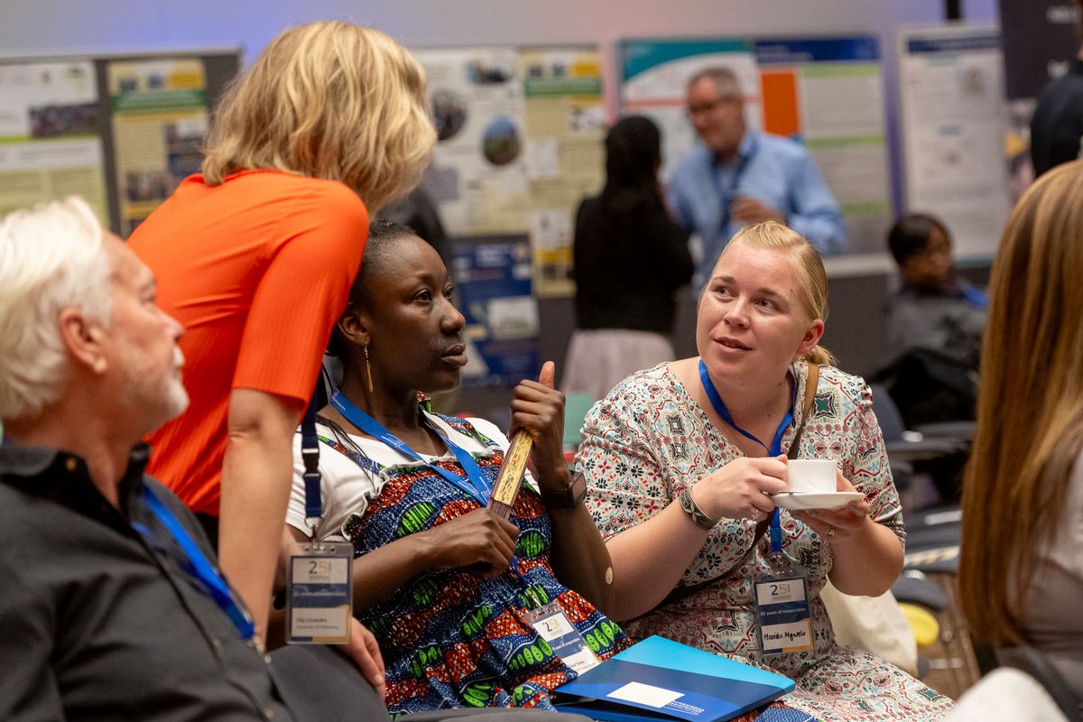 Das Bild zeigt eine Gruppe von Menschen, die in einem Raum stehen und sich unterhalten. Im Vordergrund sitzen drei Frauen, die sich intensiv unterhalten. Eine Frau trägt ein orangefarbenes Oberteil, eine andere ein bunt gemustertes Kleid und die dritte ein Oberteil mit einem floralen Muster. Alle tragen Namensschilder mit der Aufschrift "25 Years Partnership" und den Logos der Universitäten. Im Hintergrund sind weitere Personen zu sehen, die sich ebenfalls unterhalten und Poster-Aufsteller zu sehen. 