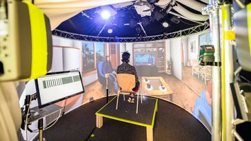 A test person sits on a chair, in the foreground some technical devices (loudspeakers, projectors) can be seen, in the background a living room scene is projected on a screen.