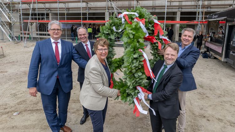 Five men in suits and a woman stand in front of the scaffolded shell and hold the topping-out wreath.