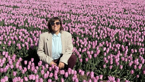 Eine junge Frau mit Sonnenbrille, Hemd und Sakko hockt in einem Feld mit blühenden rosa Tulpen und lächelt in die Kamera. 