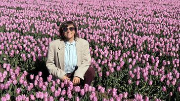 Eine junge Frau mit Sonnenbrille, Hemd und Sakko hockt in einem Feld mit blühenden rosa Tulpen und lächelt in die Kamera. 