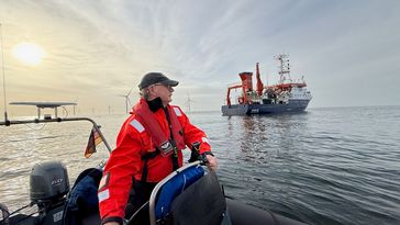 Ein Mann in Schwimmweste, orangefarbener Spezialjacke und  mit einer Schirmmütze auf dem Kopf steht am Steuer eines größeren Schlauchbootes. Das Wasser ist relativ glatt, im Hintergrund rechts das Forschungsschiff Heincke, links ein paar etwas weiter entfernte WIndräder und die untergehende Sonne, die die Szenerie in ein goldenes Licht taucht.