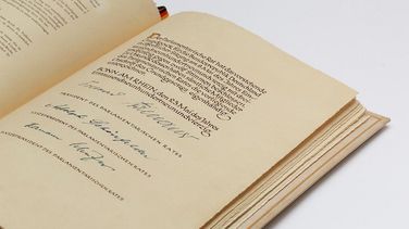 Das Bild zeigt ein aufgeklapptes Buch. Es ist die letzte Seite des Grundgesetzes mit den drei Unterschriften. Das Lesezeichen ist ein Bändchen in den Farben Schwarz-Rot-Gold.