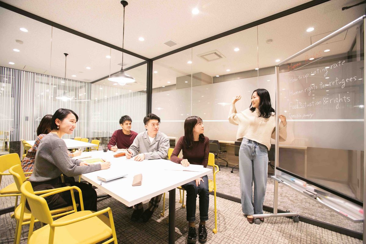 Eine Gruppe Studierender diskutiert an einem Tisch, während eine Person etwas an einem Whiteboard erklärt.