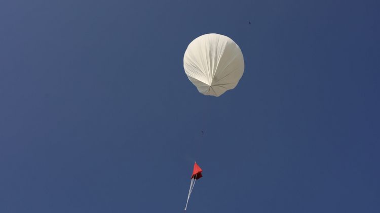 ein weißer Ballon mit dünner Schnur und rotem Fallschirm. Im Hintergrund blauer Himmel.