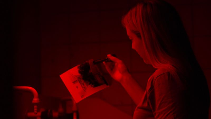 Frau in der Dunkelkammer, in ihrer Hand eine Pinzette, mit der sie ein frisch entwickeltes Foto hält.