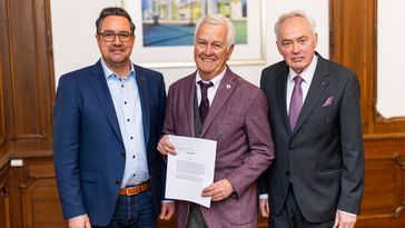 Three men in suits stand next to each other in a room with wood panelling, the middle one holding a piece of paper towards the camera.