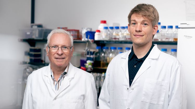 Two men in white coats in front of a laboratory shelf. 