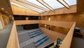 View of the interior of the building from the upper gallery. Light comes in through the roof, everything inside is clad in wood and there are landings next to the stairs where you can sit down. 