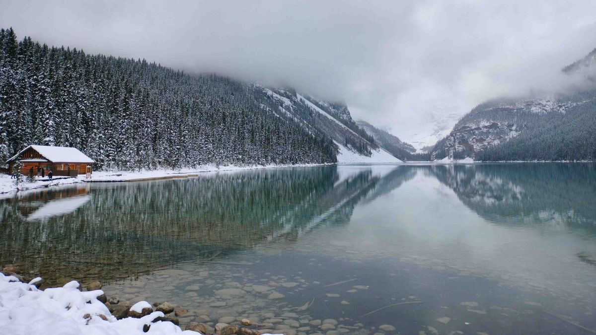Winterliche Berglandschaft mit See, verschneiten Wäldern und Hütte am Ufer.