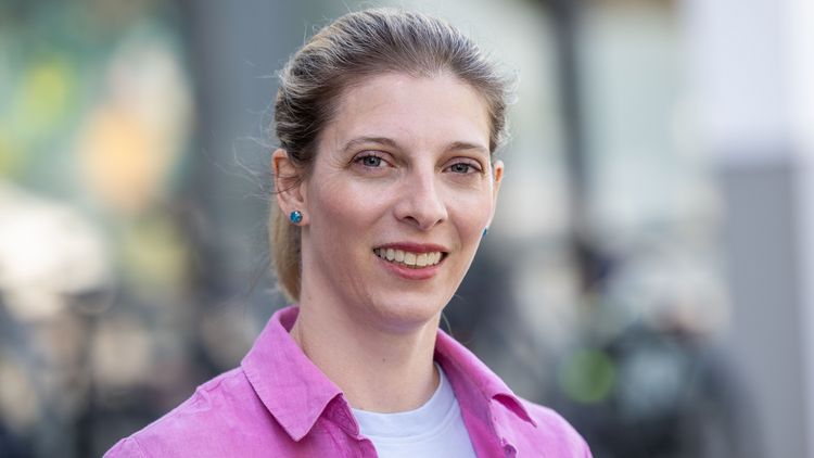 Das Bild zeigt Teresa Sansour. Sie steht vor einem Gebäude der Uni und lächelt in die Kamera.