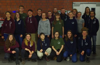 The photo shows the Environmental Sciences Student Council for winter & summer semester 2019/20. 21 students can be seen, all in their early to late 20s. They are all wearing colourful jumpers, jackets or T-shirts with the Student Council logo.  They are standing and kneeling in 3 rows. The photo was taken in the ICBM. In the background is a brick wall, on the left is a model ship in a display case, on the right is the door to a laboratory.