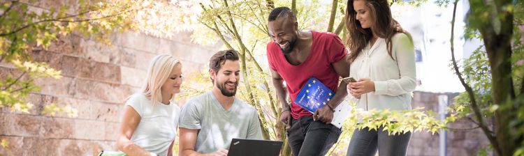Das Bild zeigt vier junge Menschen, die sich im Freien unter einem Baum befinden. Alle vier lächeln, während eine Person vor einem Laptop sitzt und tippt. Eine Person hält ein Heft mit der Flagge der Europäischen Union in den Händen, was auf ein internationales oder europäisches Projekt hindeutet. 