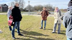 A group of people are standing on a meadow, with the building site in the background.