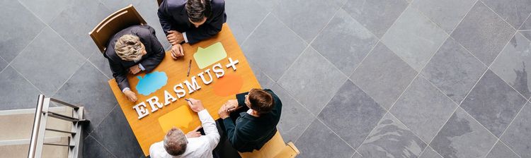 Vier Personen sitzen an einem Tisch. Auf dem Tisch steht der Schriftzug "Erasmus+". 