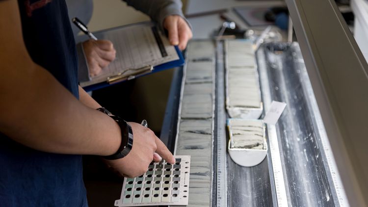 The picture shows a core sample that has been cut open and split in half, sitting inside a metal cylinder. Two researchers are standing next to it; one is writing something on a clipboard, whilst the other is pointing at a colour chart.