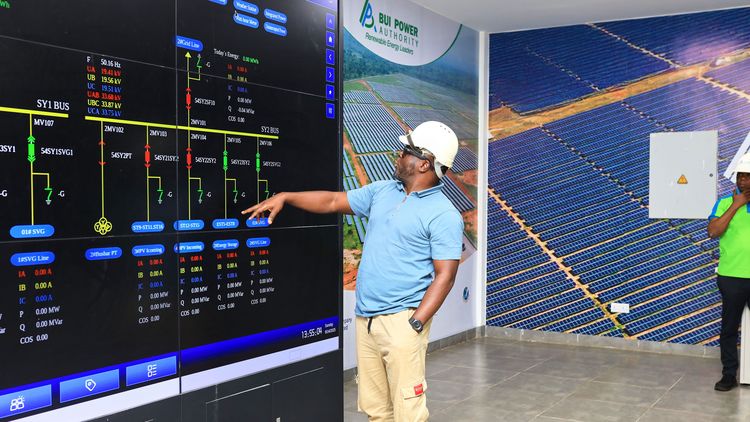 Ein Mann mit Helm und Schutzbrille steht vor einem riesigen Bildschirm, auf dem Schaubilder und Zahlen zu sehen sind. Die Wände im Hintergrund sind von Fotos riesiger Solarparks bedeckt. 