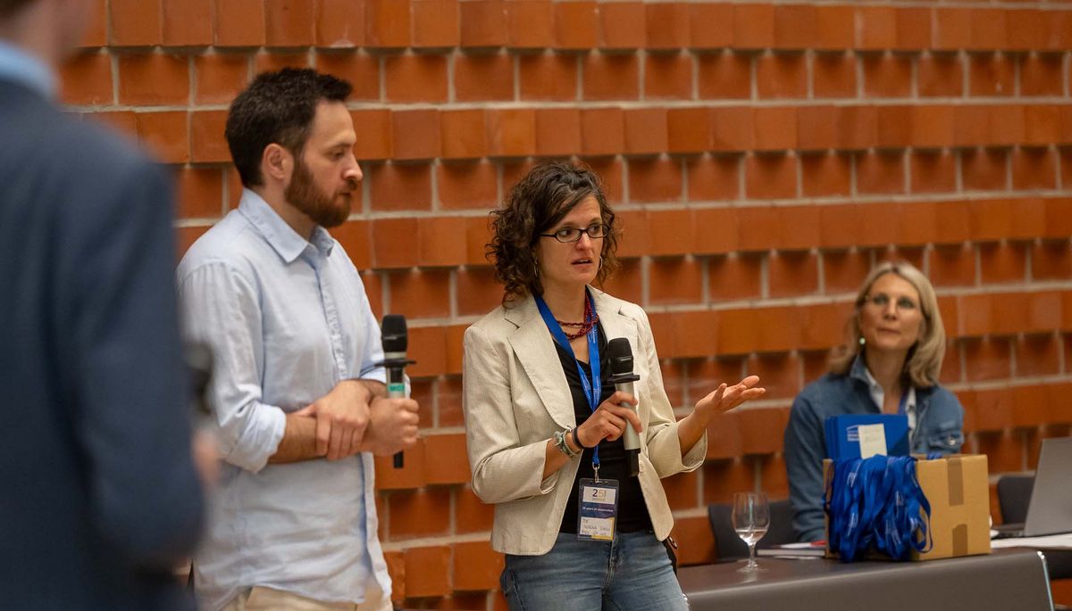 Zwei Personen stehen vor einer roten Backsteinwand und sprechen in Mikrofone. Eine Frau in einem beigen Blazer gestikuliert lebhaft, während ein Mann neben ihr mit einem Mikrofon in der Hand zuzuhören scheint. Im Hintergrund ist eine weitere Person zu sehen. Das Bild vermittelt den Eindruck einer Diskussion oder einer Fragerunde während des Symposiums. Beide Personen scheinen aktiv an der Veranstaltung teilzunehmen.