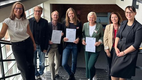 Das Bild zeigt die genannten Personen stehend auf einer Treppe. Alle lächeln in die Kamera, die drei Geprüften halten ihre Urkunden in den Händen.