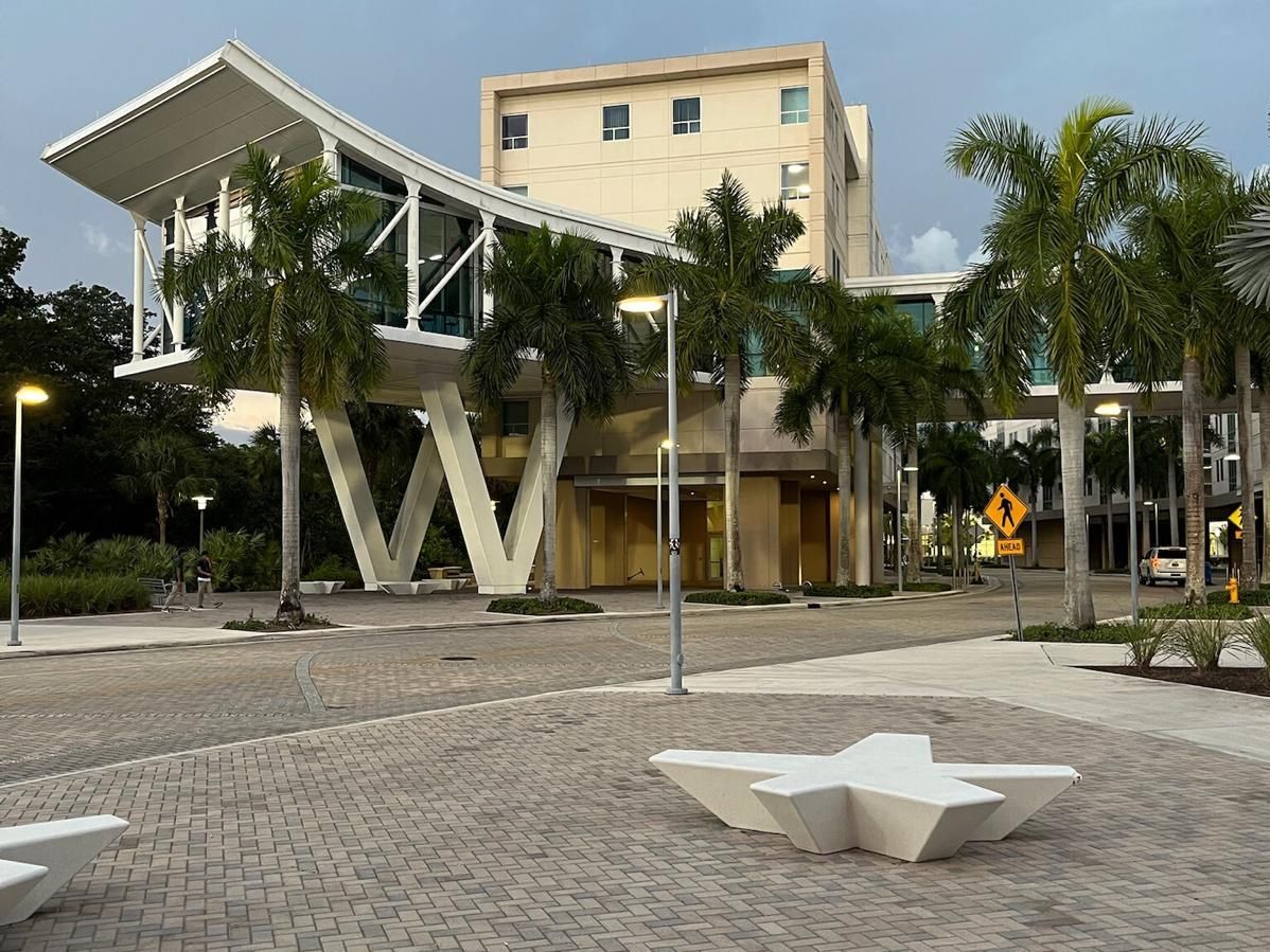 Modernes Gebäude mit überdachtem Übergang, Palmen und gepflastertem Platz davor.