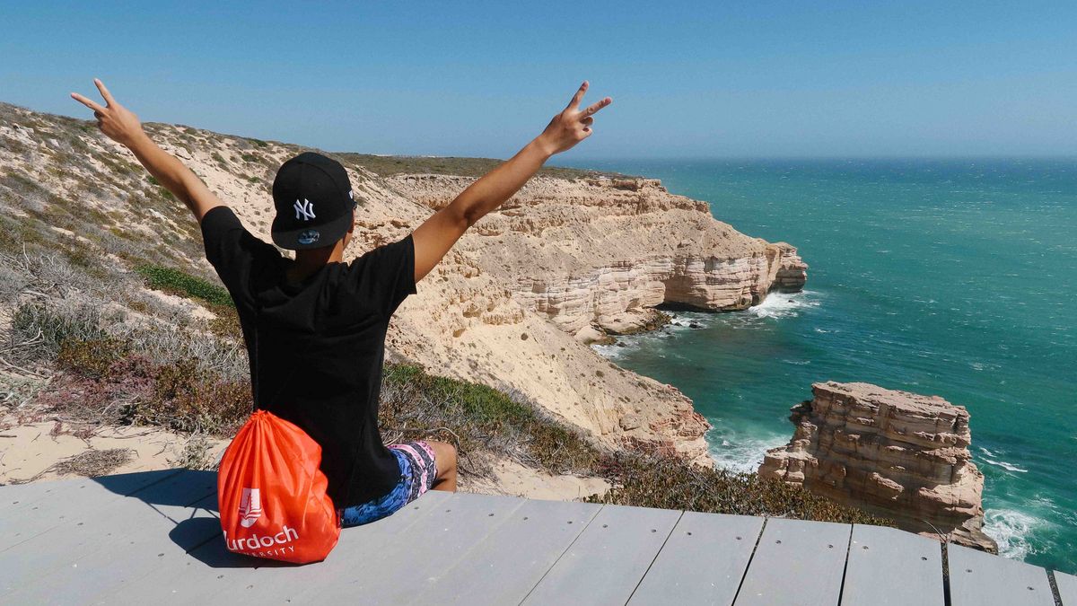 Eine Person steht mit ausgebreiteten Armen auf einer Holzplattform an einer Klippe und blickt aufs Meer. Eine Murdoch University Tragetasche liegt in der Nähe.