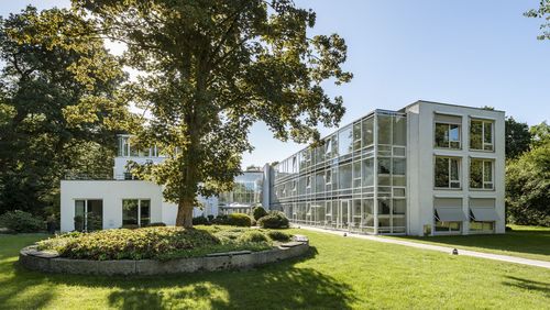 Blick auf das weiße Gebäude, umgeben von einer parkartigen Landschaft,  von der Rückseite aus.