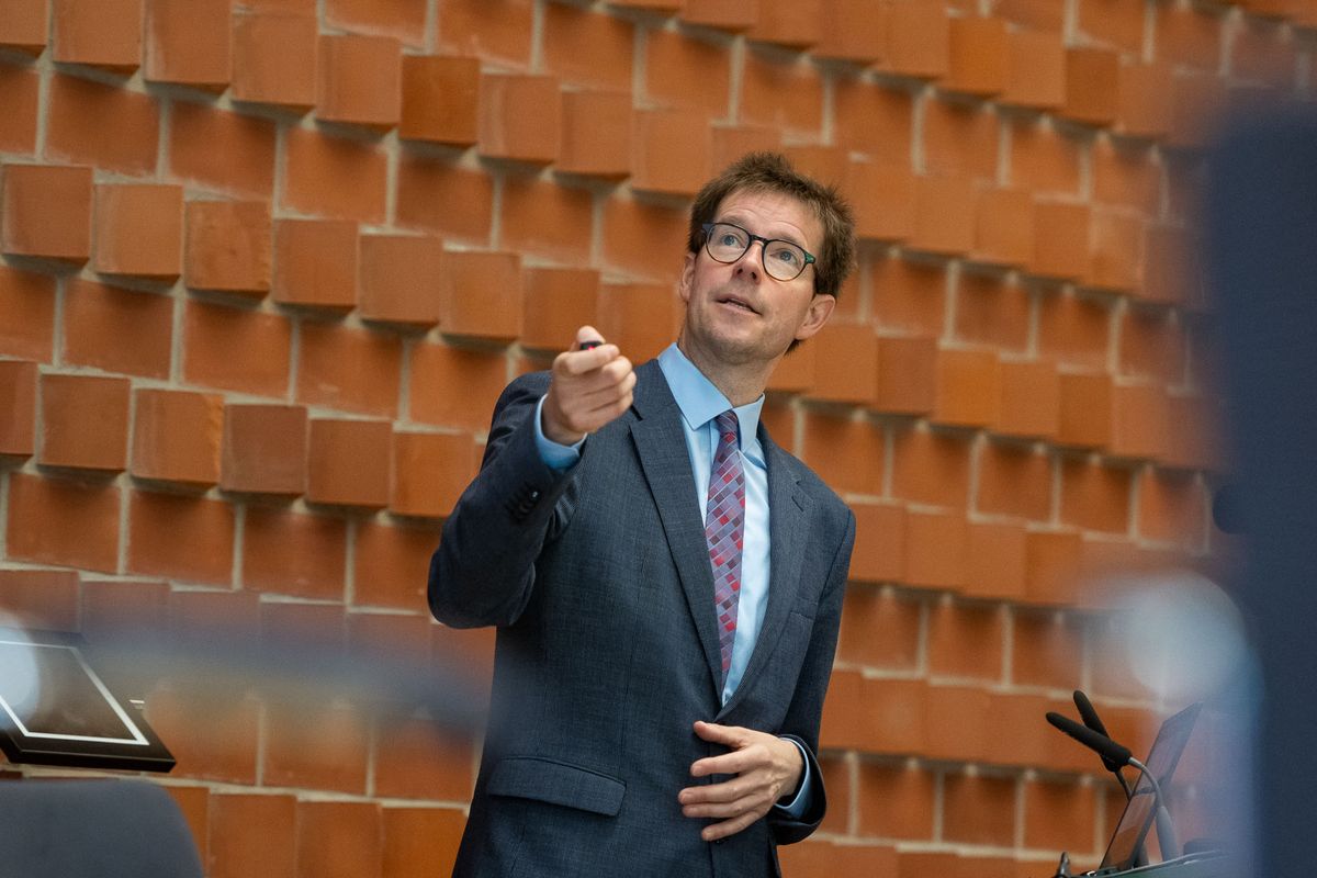 Ein Mann in einem grauen Anzug mit bunter Krawatte steht vor einer roten Backsteinwand und hält eine Rede. Er trägt eine Brille und deutet mit der rechten Hand auf etwas außerhalb des Bildes. Ein Mikrofon steht vor ihm auf einem Pult.