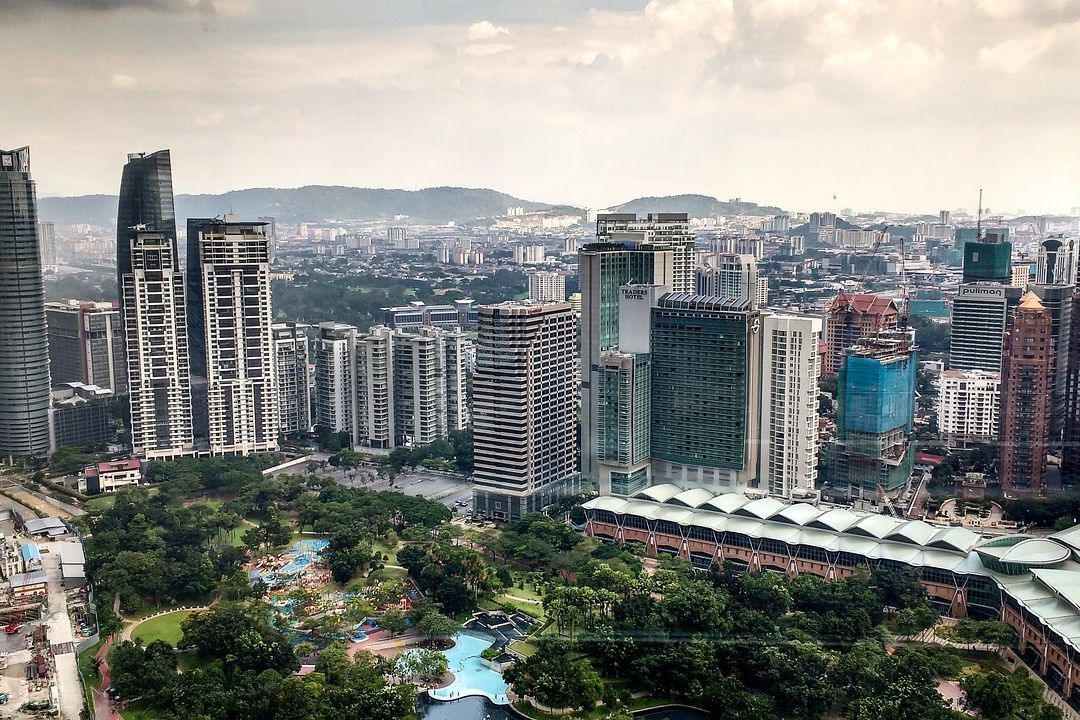 Ein Panorama der Stadt Kuala Lumpur, Malaysia, mit hohen Wolkenkratzern. Im Vordergrund ein Park mit Bäumen und Schwimmbad.