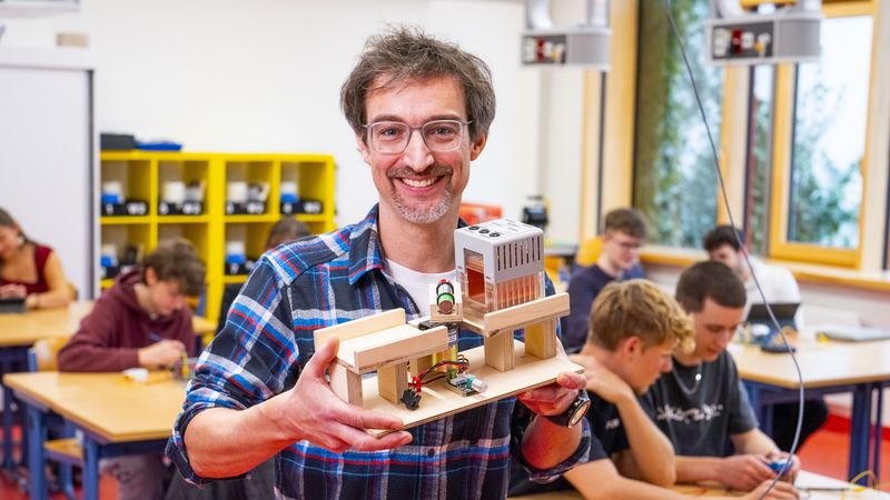 Ein Mann hält ein Holzmodell in der Hand, im Hintergrund ein Klassenzimmer, in dem Schülerinnen und Schüler elektronische Bauteile ausprobieren. 