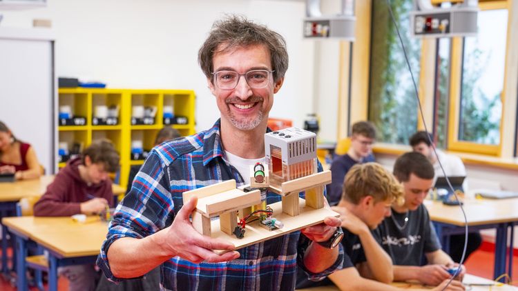 Ein Mann hält ein Holzmodell in der Hand, im Hintergrund ein Klassenzimmer, in dem Schülerinnen und Schüler elektronische Bauteile ausprobieren. 