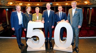 Sechs Personen stehen hinter einer großen 50 auf der Bühne des Staatstheaters, im Hintergrund der leere Publikumsbereich des Theaters. 