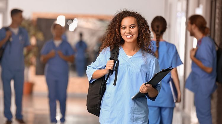 A young woman in work clothes is in a medical facility. Other colleagues can be seen in the background.