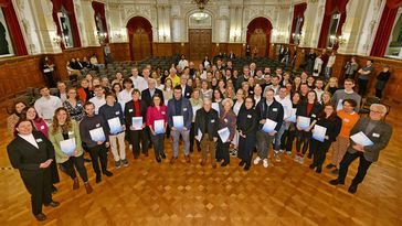 The picture shows the recipients and their sponsors in a large group photo. They look into the camera and show their certificates. The photo was taken in the magnificent ballroom of Oldenburg Palace. It features stucco-decorated walls and ceilings, chandeliers and red curtains.