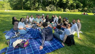 Das Bild zeigt eine Gruppe von Menschen, die in einem Park auf dem Rasen sitzen und ein Picknick veranstalten. Im Hintergrund sind Büsche zu erkennen, die Sonne scheint.