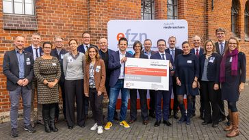A large group of people are standing in front of a red brick building, two men are holding a symbolic cheque for 58 million euros.
