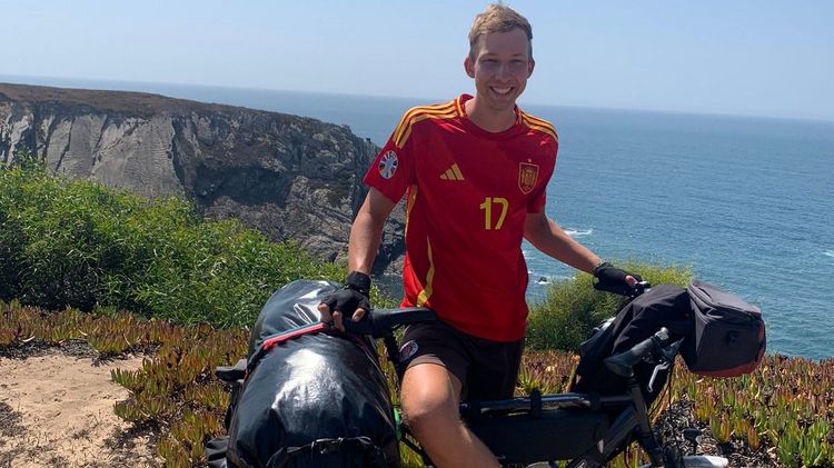 Jasper Laschinger steht an seinem Rad und lacht in die Kamera. Er trägt ein Trikot der spanischen Fußballnationalmannschaft (Nr. 17). Im Hintergrund sind eine Steilküste, blauer Himmel und das Meer zu erkennen.