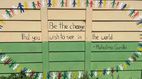 Eine bemalte Wand mit dem Zitat von Mahatma Gandhi: "Be the change that you wish to see in the world". Darüber und darunter sind bunte, stilisierte Figuren von Menschen gemalt.
