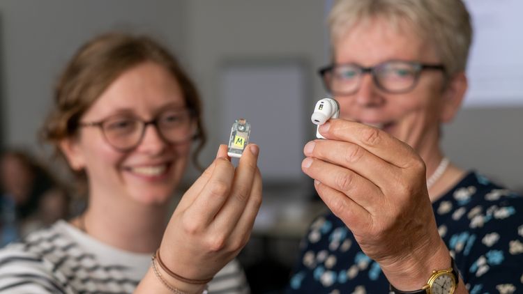 A young and a slightly older woman are each holding a small device up to the camera that looks like an in-ear headphone.