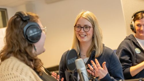 Zwei Frauen mit großen Kopfhörern und einem Mikrofon, eine weitere Frau erklärt die Funktionsweise. eine 