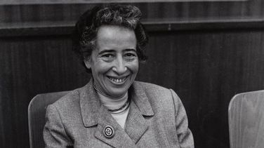 The picture shows a black-and-white photograph of Hannah Arendt. She is sitting on a chair and smiling warmly at the camera.